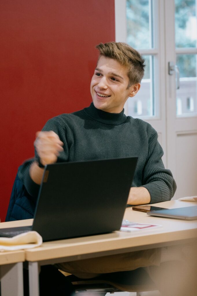 Junger Mann in einem grünen Shirt schaut nach links und am sprechen. Hintergrund rote Hand und Fenster.