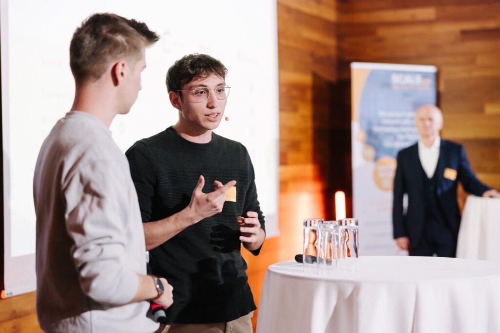 Junger Mann mit Mikrofon schaut auf die Seite und spricht. Dunkles Shirt an einem weißen Tisch stehen. Öffentliche Veranstaltung