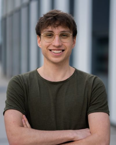 Jorrit Kappel Junger Mann in grünen T-Shirt, heller Haut, Brille und braunen Haaren schaut lachend zentral in die Kamera
